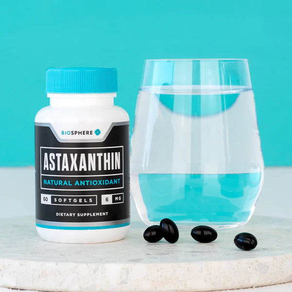A white bottle of Biosphere Astaxanthin dietary supplement with a blue cap and black label, shown next to a clear glass of water and three black softgels on a light stone surface against a turquoise background.